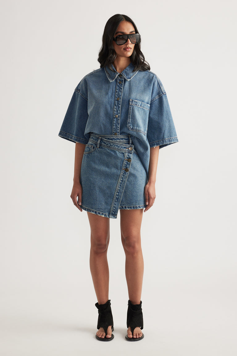 Woman wearing an asymmetric denim skirt with a matching oversized denim shirt, sunglasses and black ankle boots