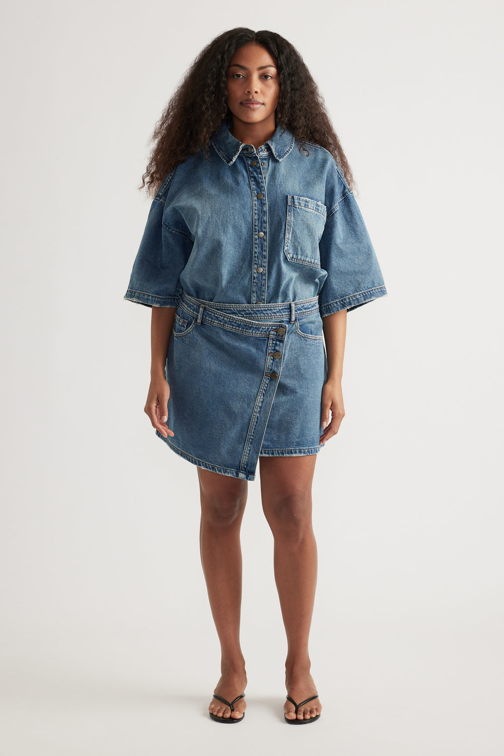 Model wearing a high rise asymmetric denim skirt with an oversized short sleeve denim shirt and flat sandals