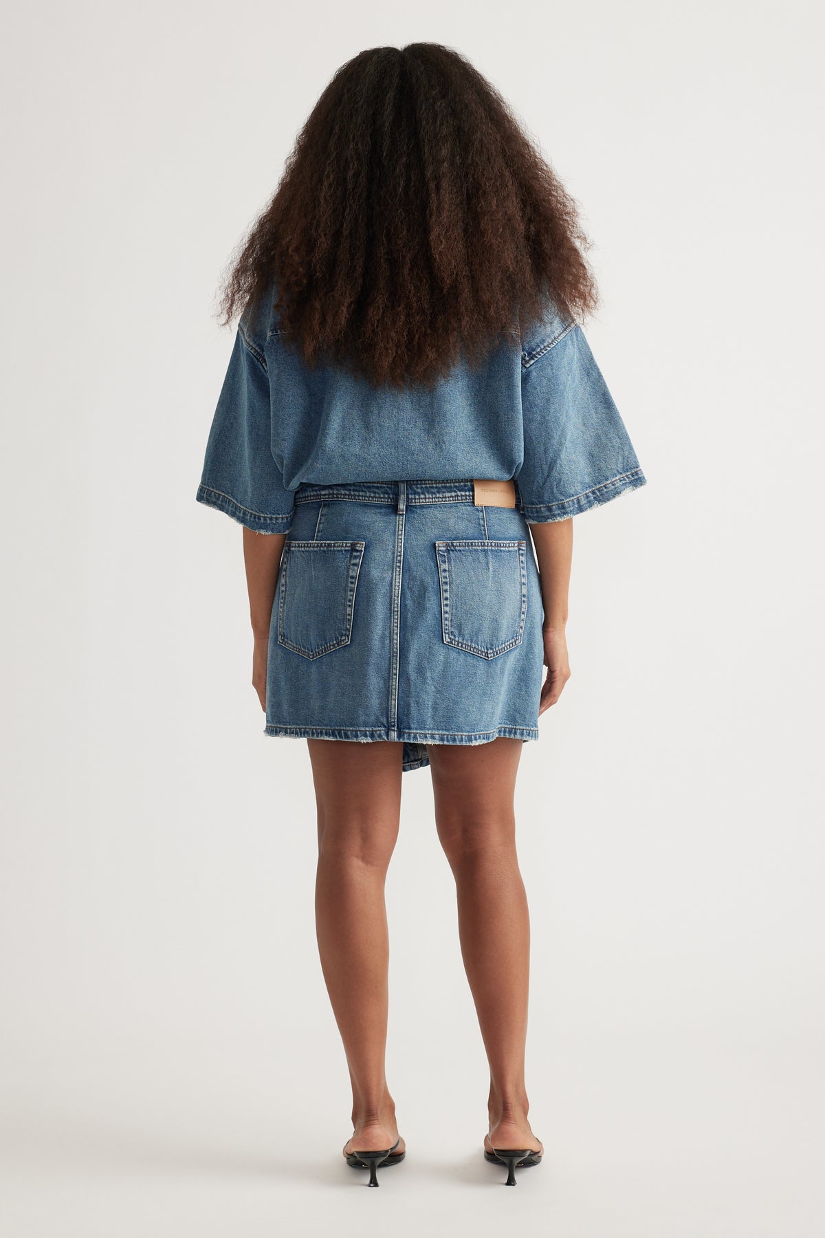 Back view of a woman wearing a denim mini skirt, cropped denim jacket and black kitten heels