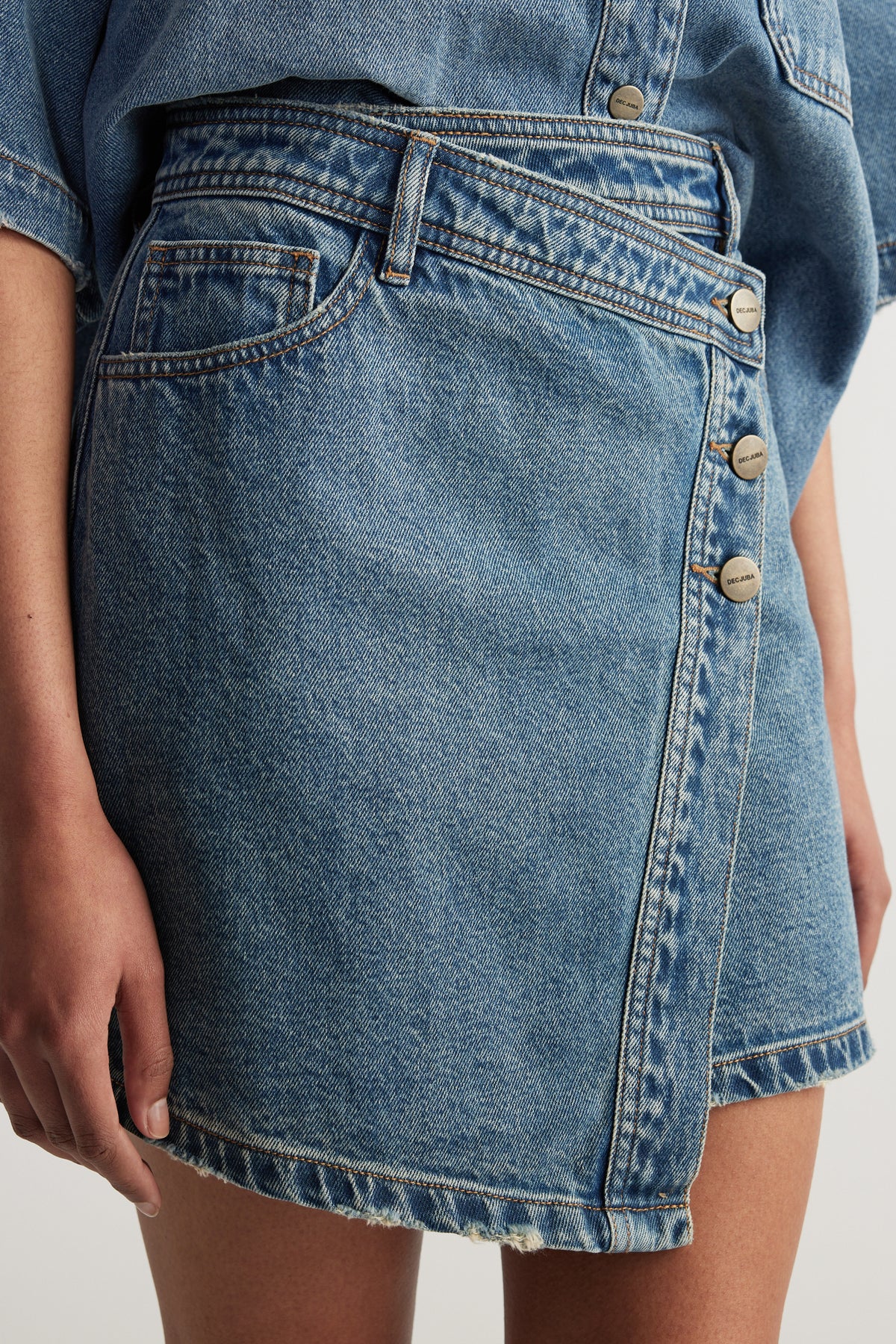 Model wearing a high rise blue denim mini skirt with an asymmetric front and button details