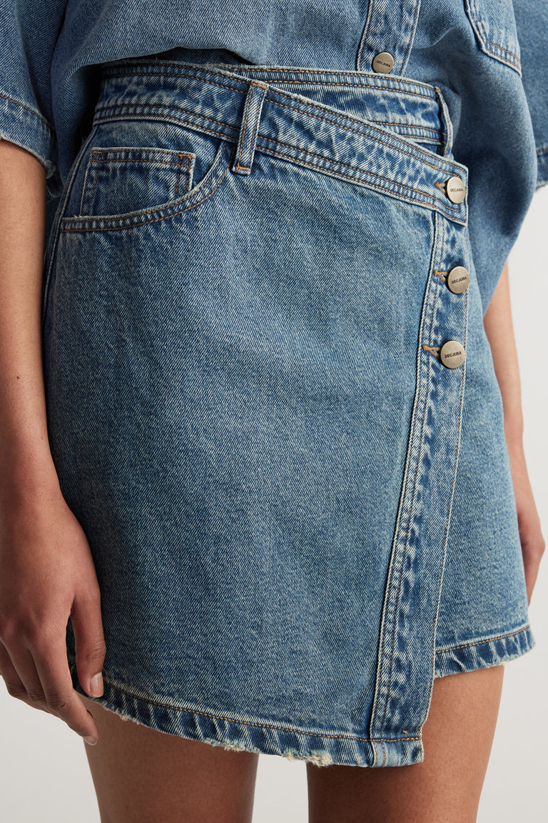 Model wearing a high rise blue denim mini skirt with an asymmetric front and button details