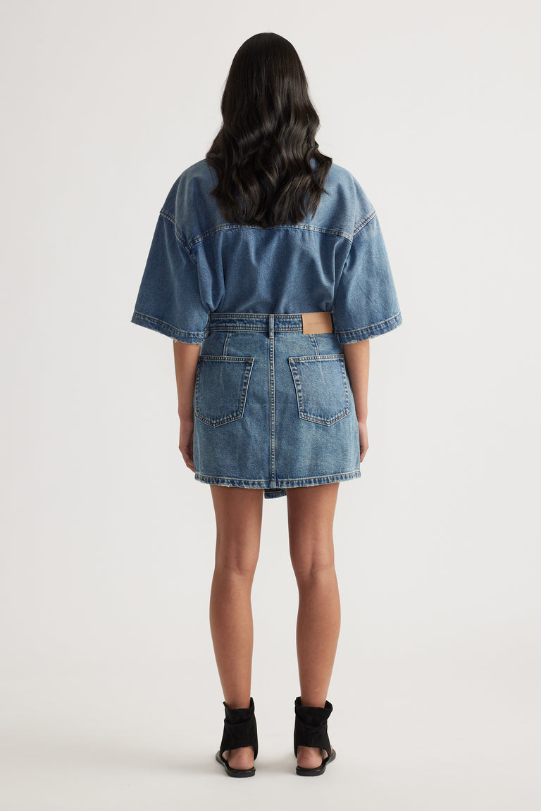Back view of a model wearing a blue denim mini skirt with patch pockets, an oversized denim top and black ankle boots
