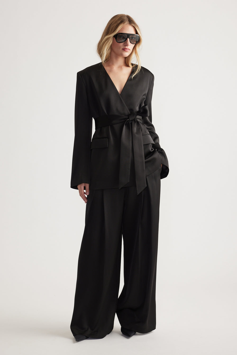 Woman wearing a black satin belted blazer with wide-leg pants and oversized sunglasses for a chic look.