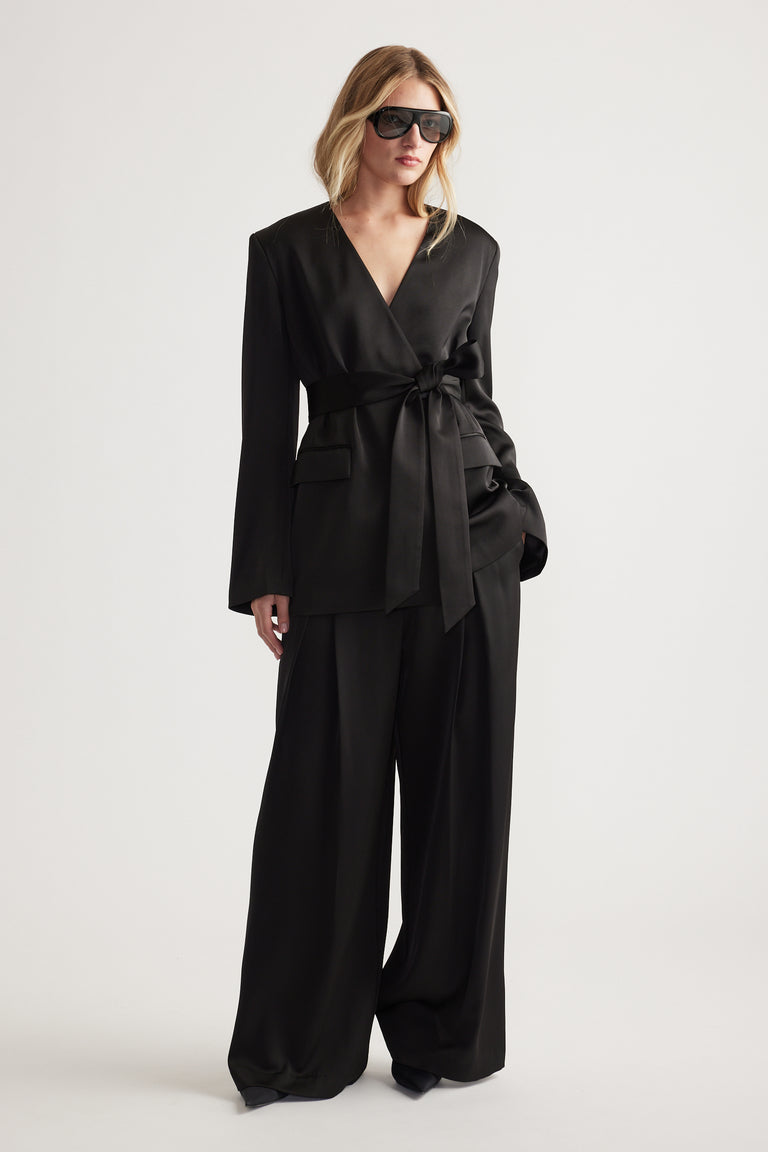 Woman wearing a black satin belted blazer with wide-leg pants and oversized sunglasses for a chic look.