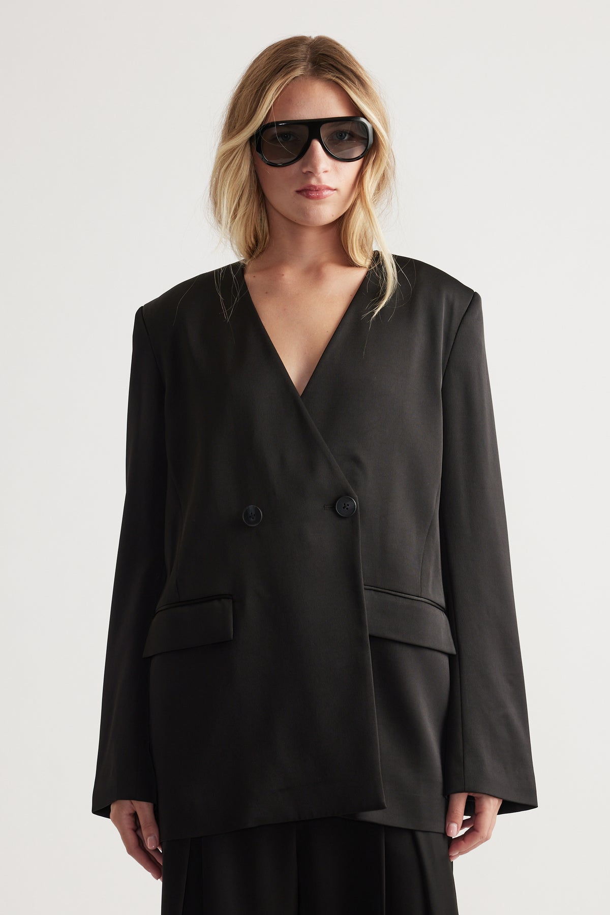 Woman wearing an oversized black satin blazer with a deep V-neckline and double-button closure paired with sunglasses.