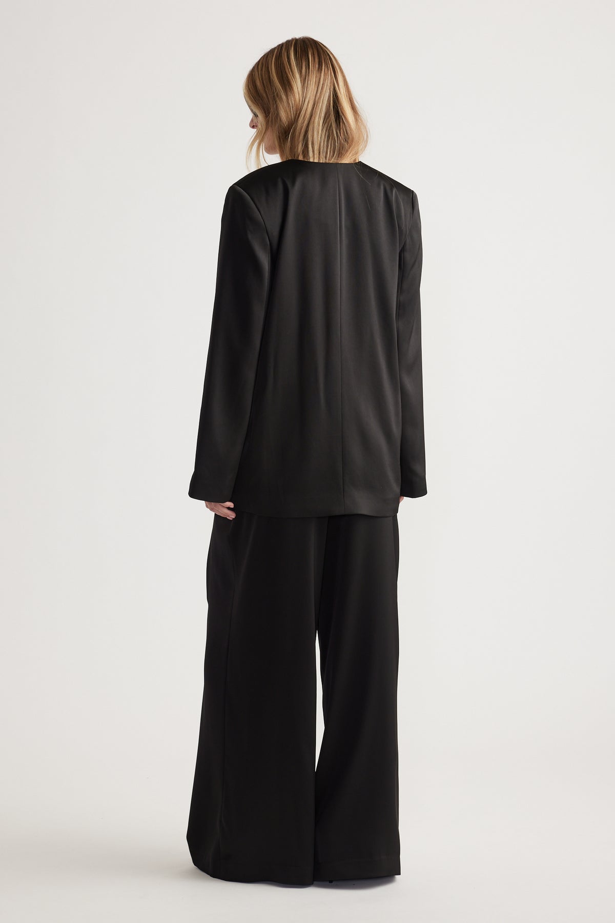 Woman with blonde hair wearing a sleek black blazer and wide-leg pants in a relaxed, elegant fit viewed from behind.