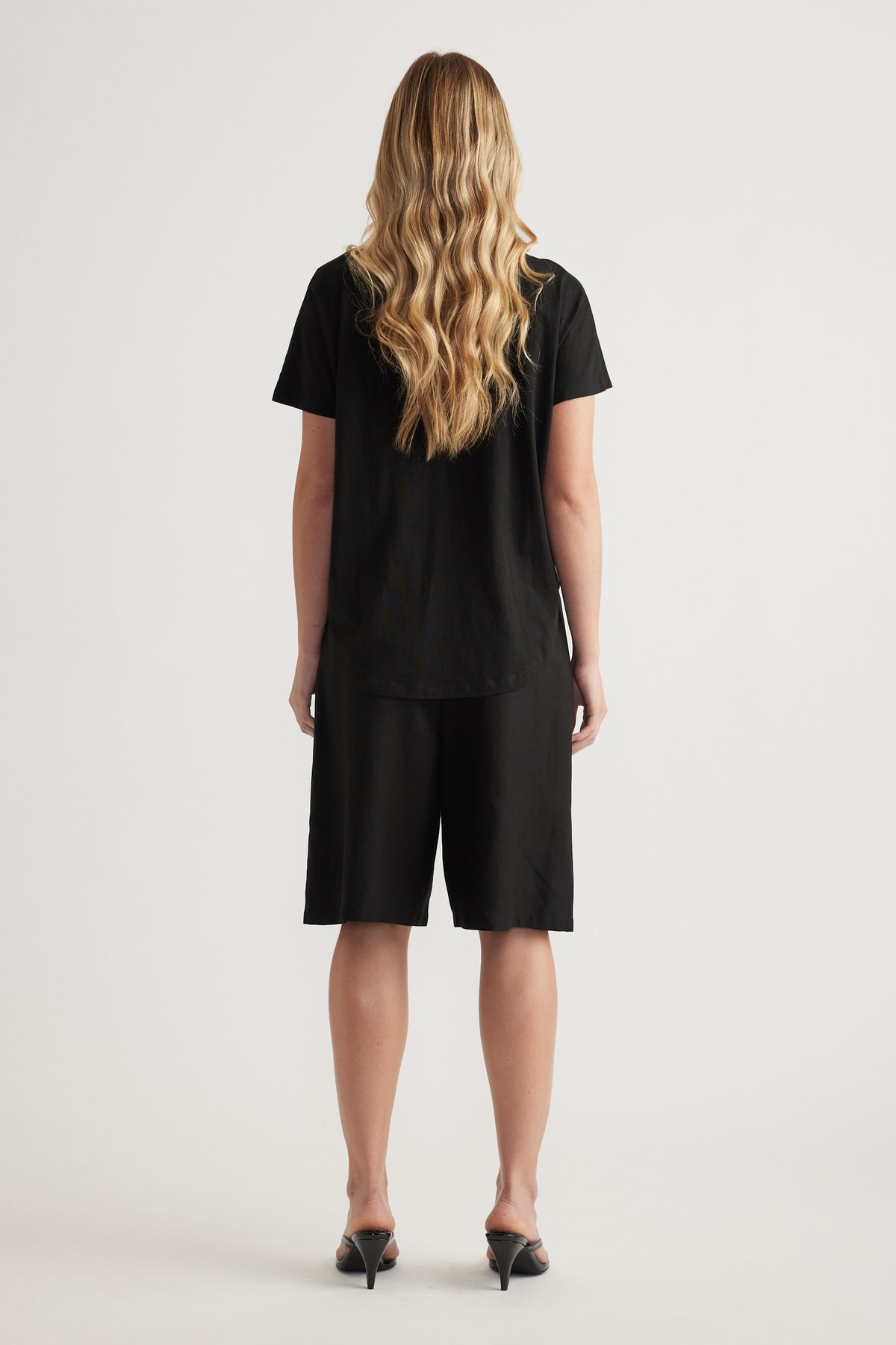 Back view of a woman wearing a black boxy tee with black knee-length shorts and heels