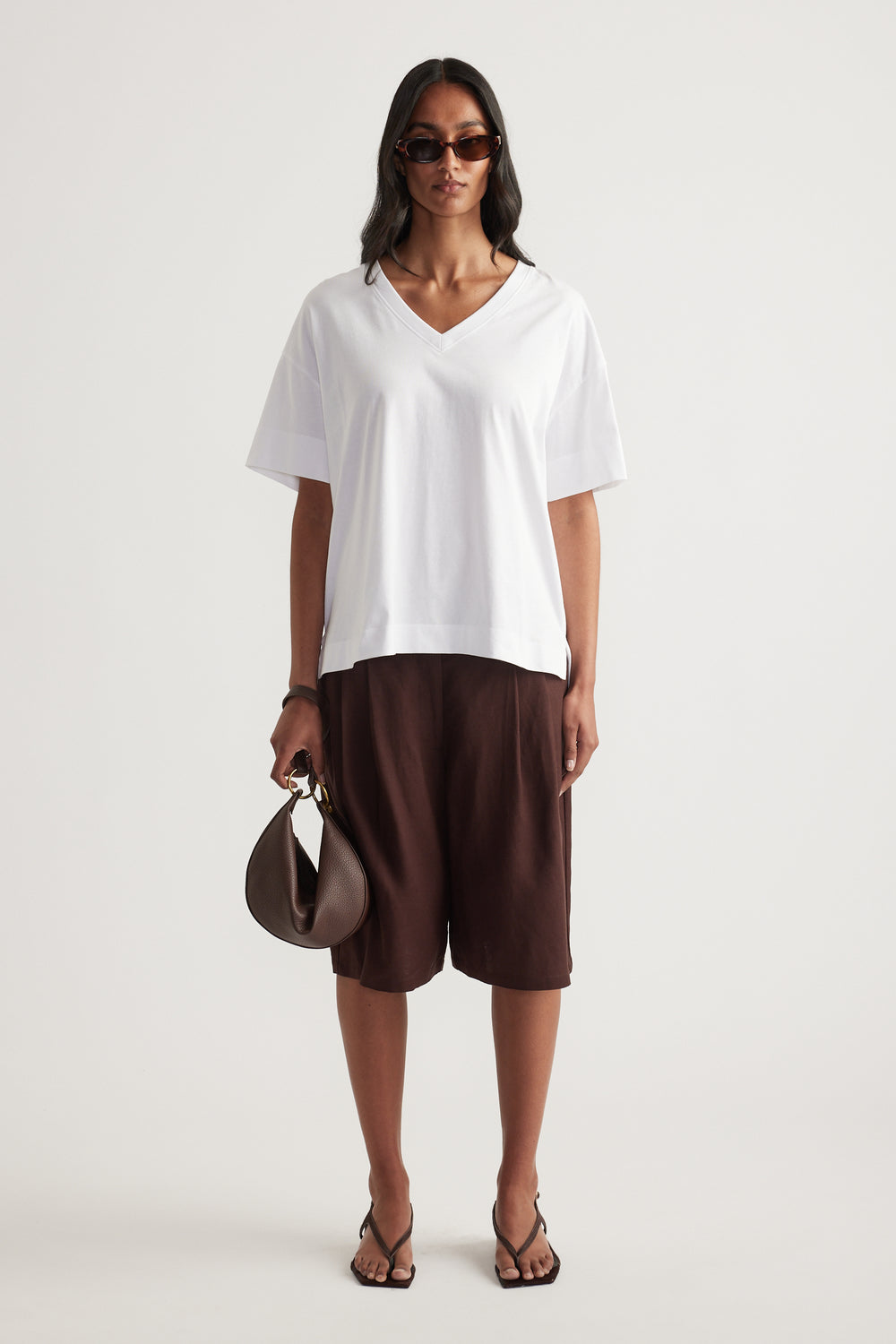 Woman wearing a white boxy V neck tee with brown shorts, sunglasses, handbag and strappy sandals