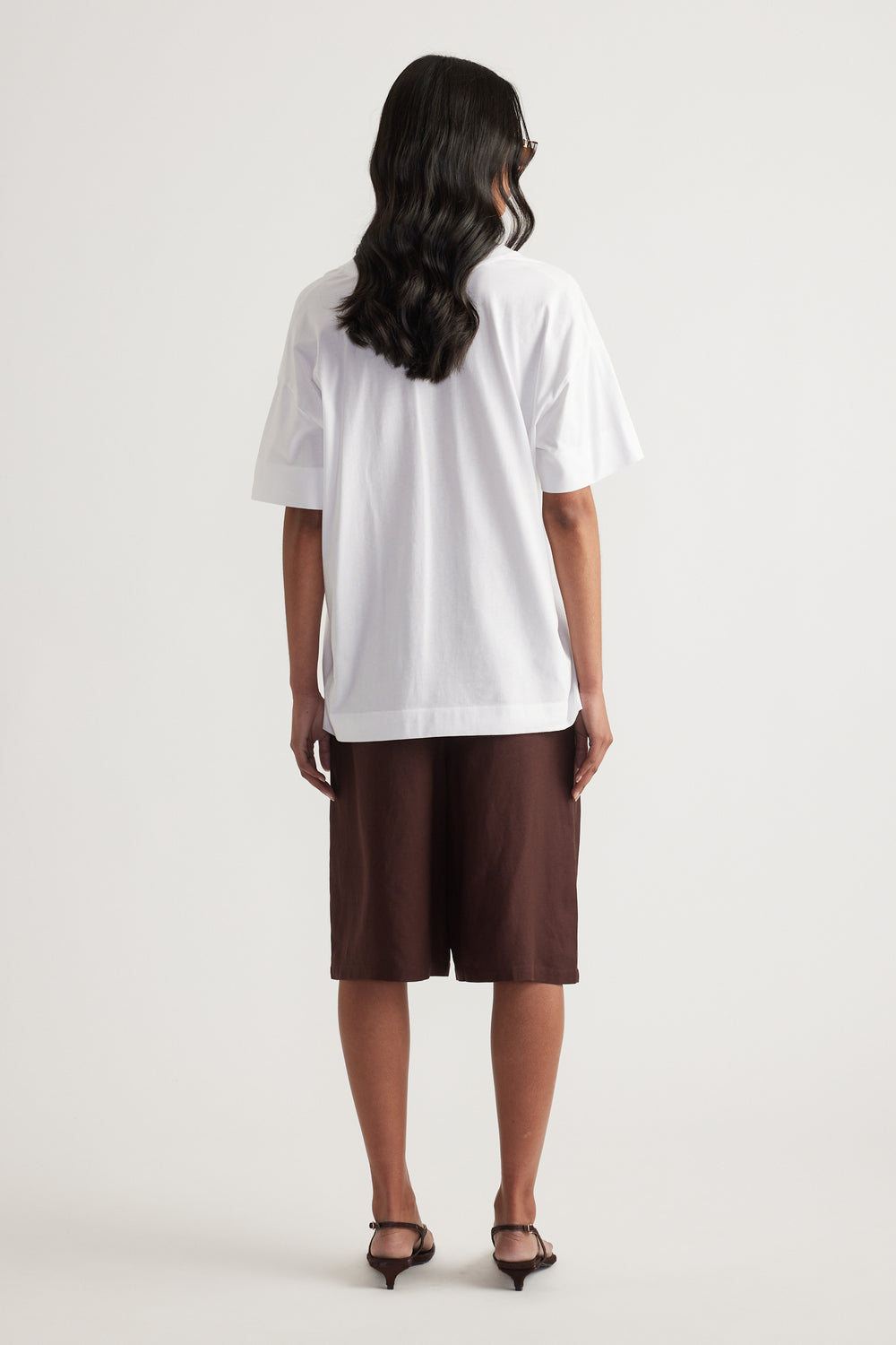 Back view of a woman wearing a white boxy tee with brown knee length shorts and black heeled sandals