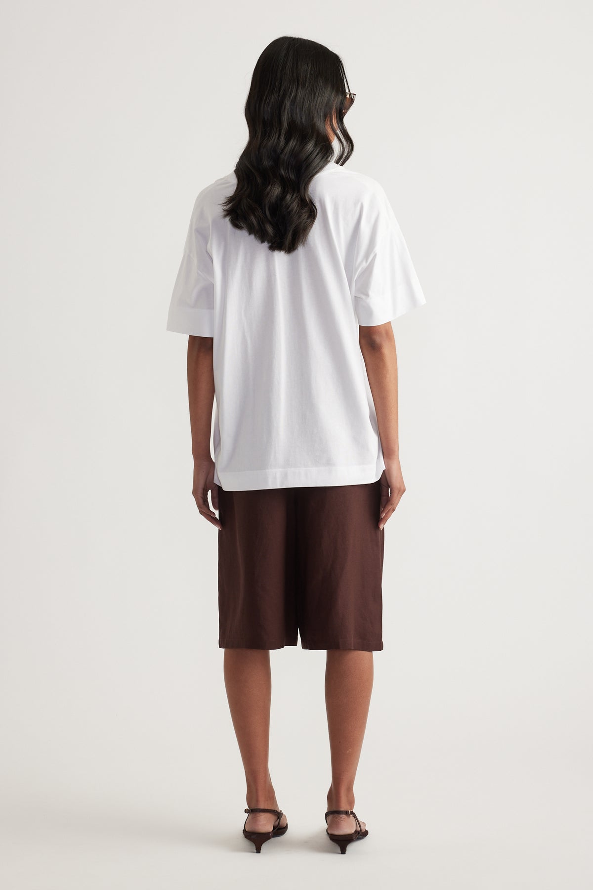 Back view of a woman wearing a white boxy tee with brown knee length shorts and black heeled sandals