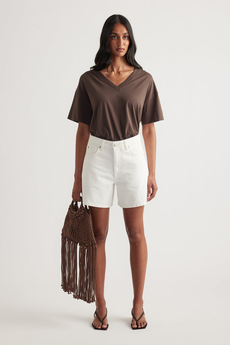 Woman wearing a brown boxy V neck tee with white denim shorts, strappy sandals and a fringed bag