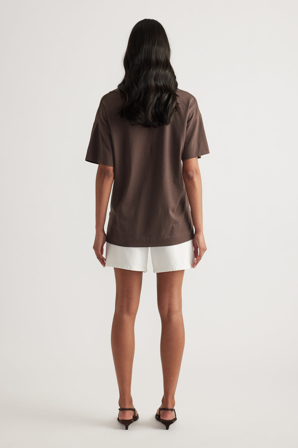 Back view of a woman wearing a brown boxy short sleeve tee, white shorts and black heels