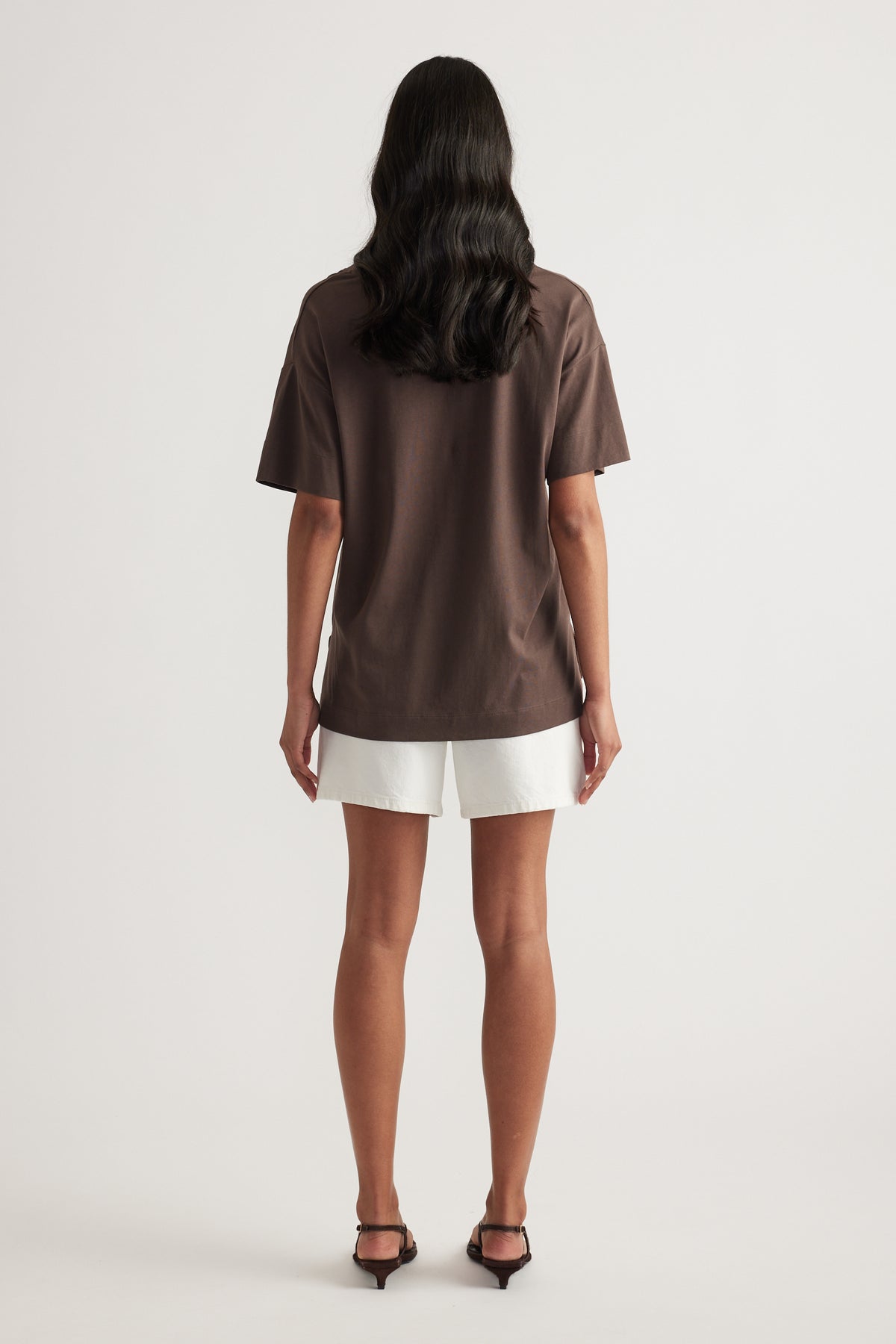 Back view of a woman wearing a brown boxy short sleeve tee, white shorts and black heels