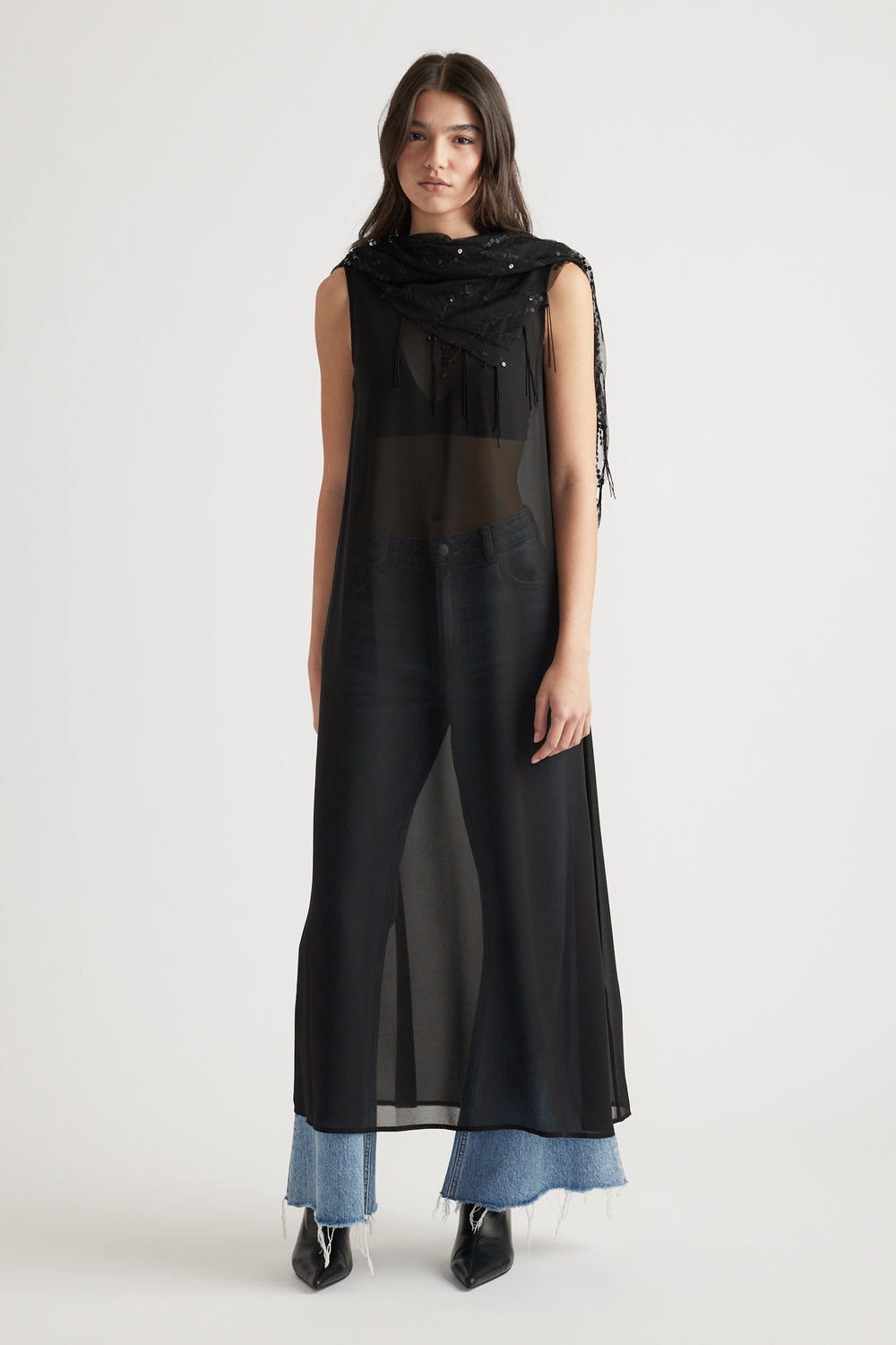 Woman wearing a sheer black maxi dress layered over blue wide-leg jeans and pointed black boots in soft light