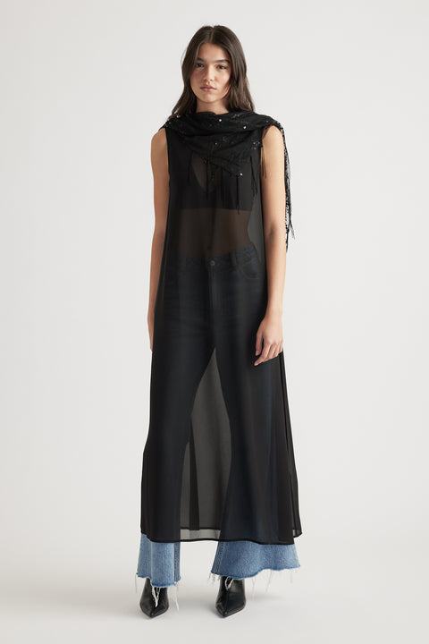 Woman wearing a sheer black maxi dress layered over blue wide-leg jeans and pointed black boots in soft light