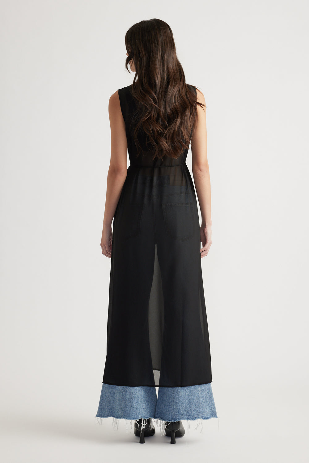 Back view of sheer black maxi dress layered over wide-leg light denim jeans with raw hems