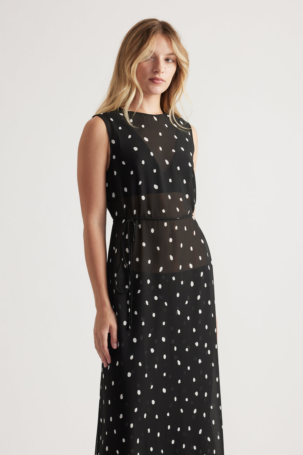 Woman wearing a sleeveless black maxi dress with white irregular polka dots and a sheer waist panel with tie detail