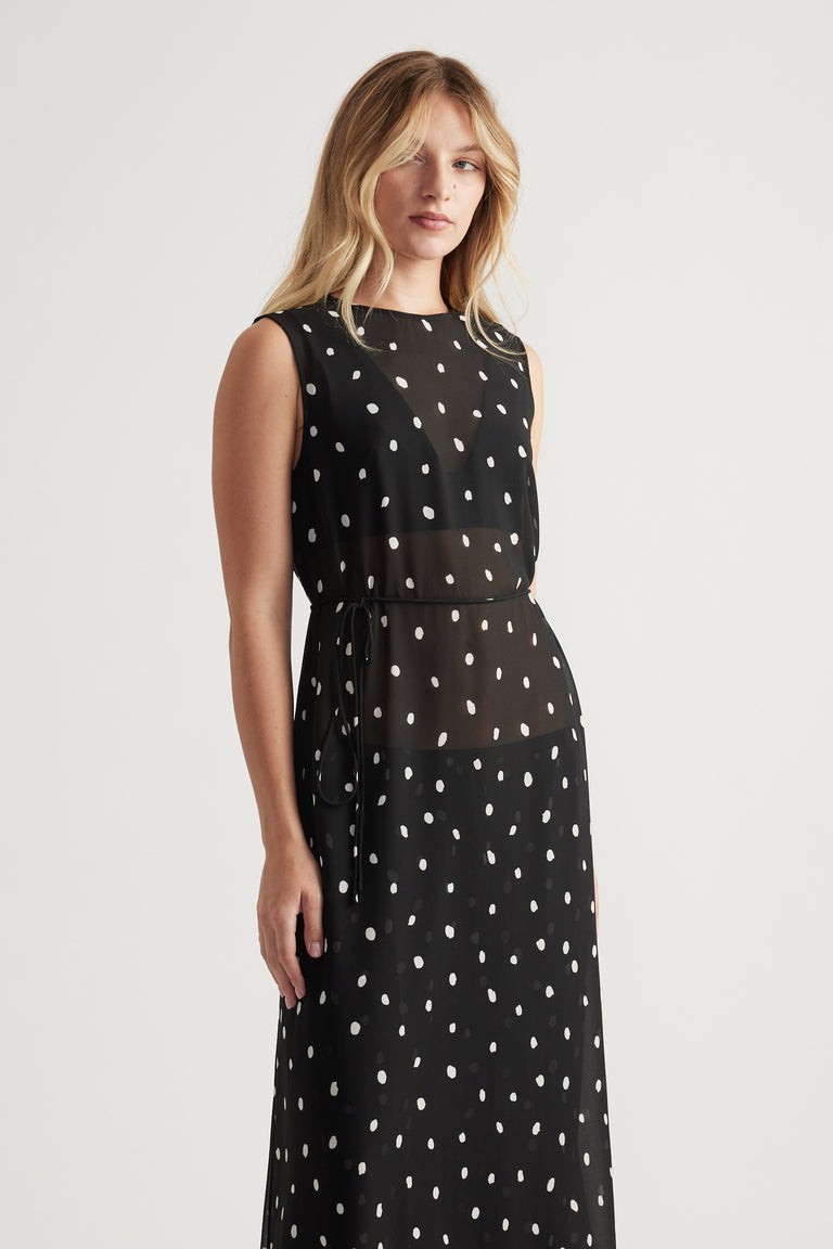 Woman wearing a sleeveless black maxi dress with white irregular polka dots and a sheer waist panel with tie detail