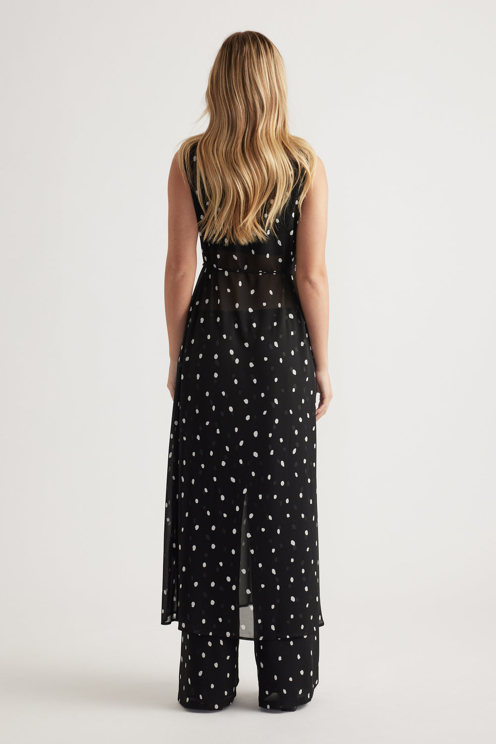 Woman wearing a black sheer maxi dress with white polka dots layered over matching wide-leg pants, back view