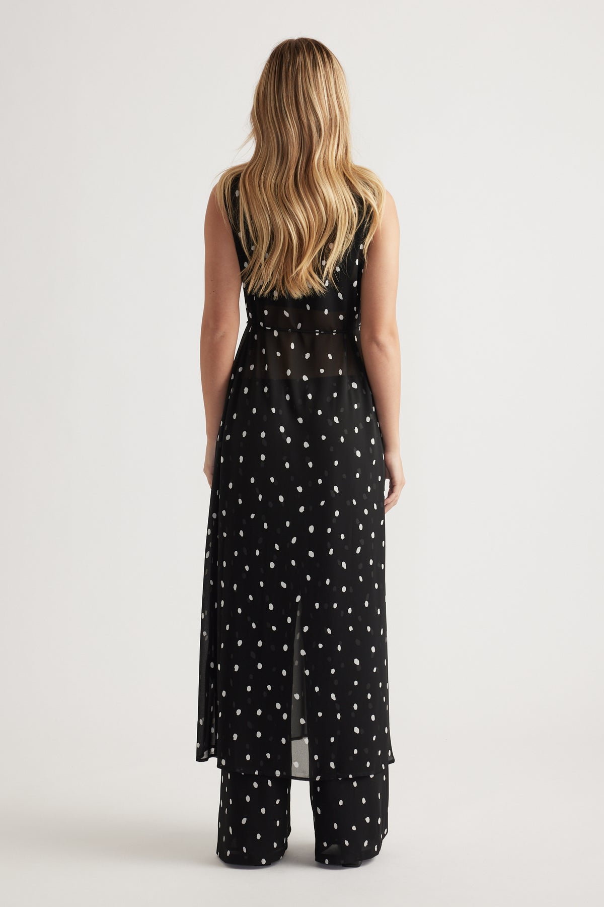 Woman wearing a black sheer maxi dress with white polka dots layered over matching wide-leg pants, back view