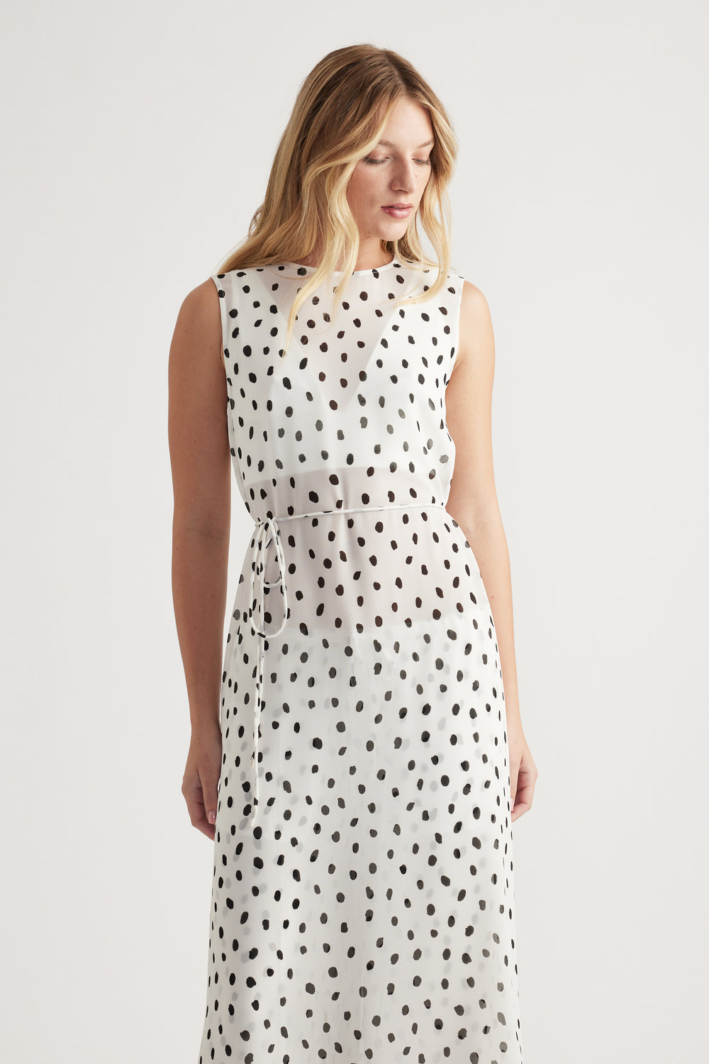 Woman wearing a sleeveless white maxi dress with black irregular polka dots and a tied waist detail