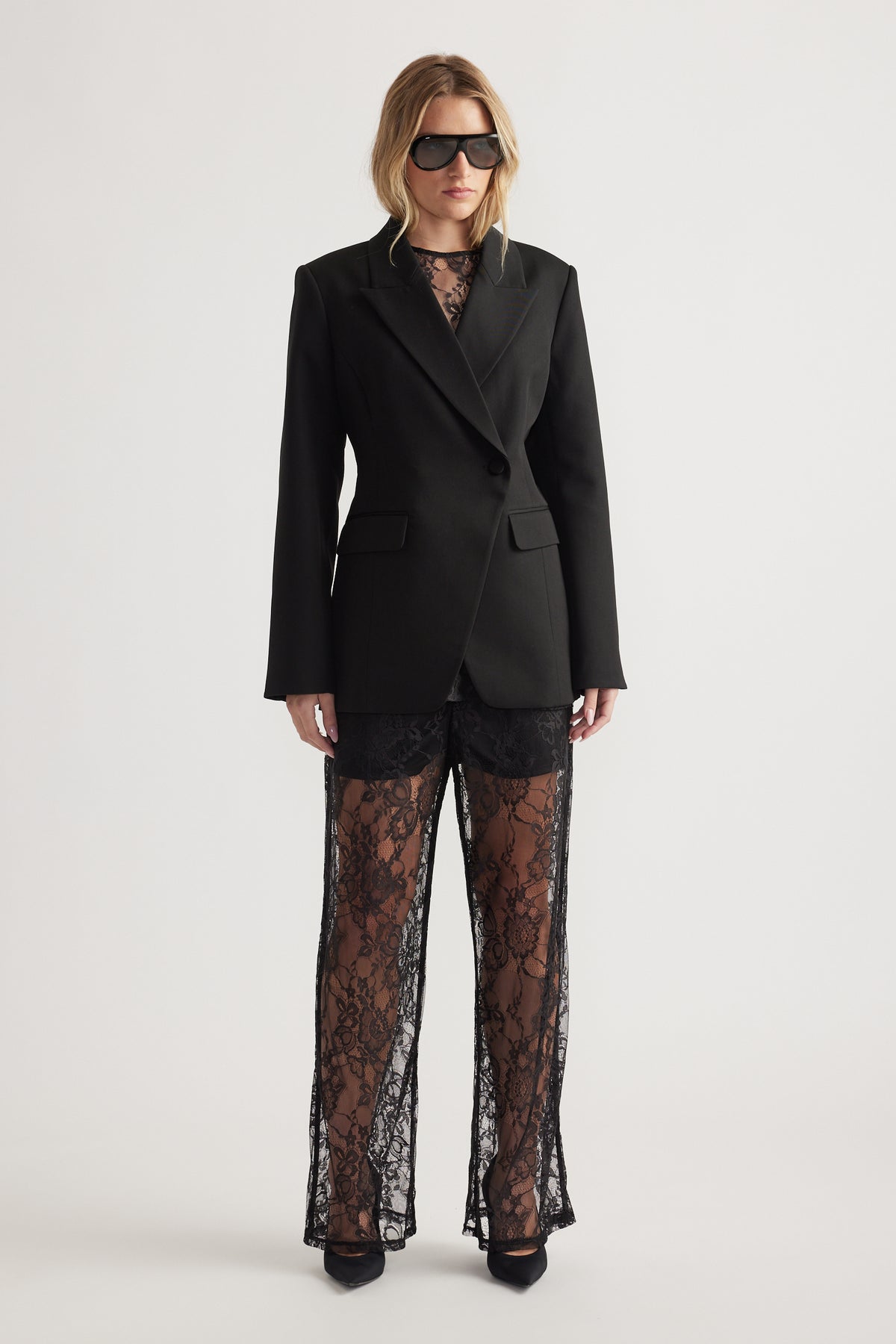 Woman wearing a tailored black blazer over sheer lace pants and pointed black heels against a plain background