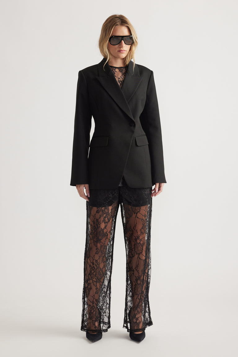 Woman wearing a tailored black blazer over sheer lace pants and pointed black heels against a plain background