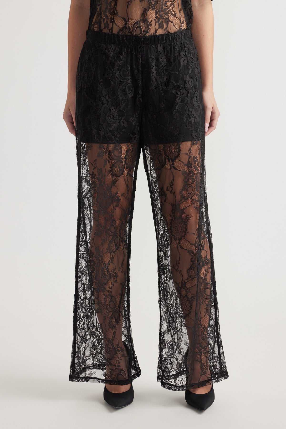 Black wide-leg lace pants with elastic waist and detachable lining worn with black heels on neutral background