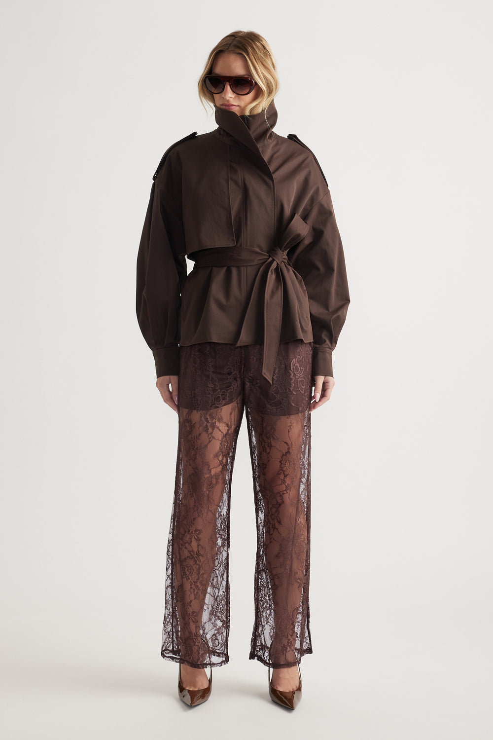 Woman wearing oversized dark brown jacket with tie waist and sheer wide-leg lace pants paired with heels