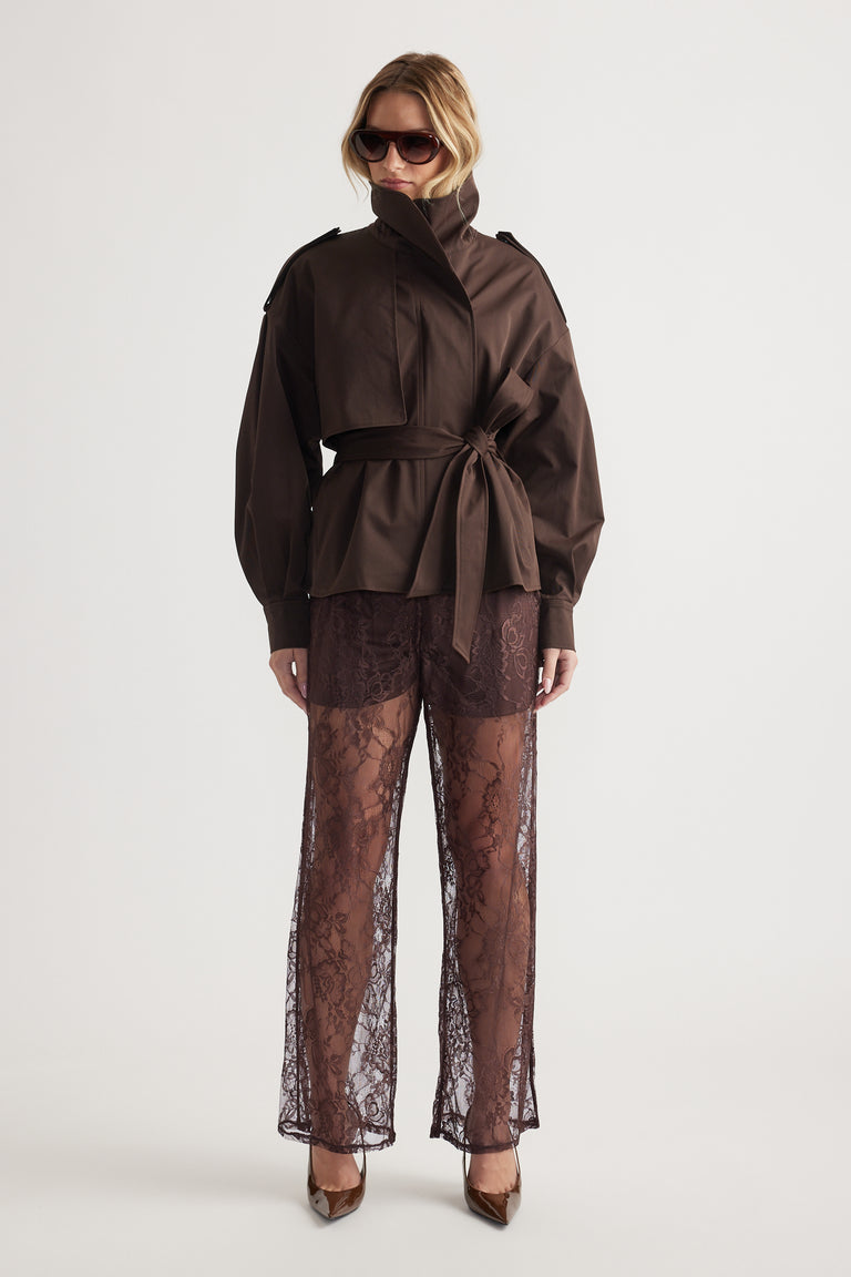 Woman wearing oversized dark brown jacket with tie waist and sheer wide-leg lace pants paired with heels