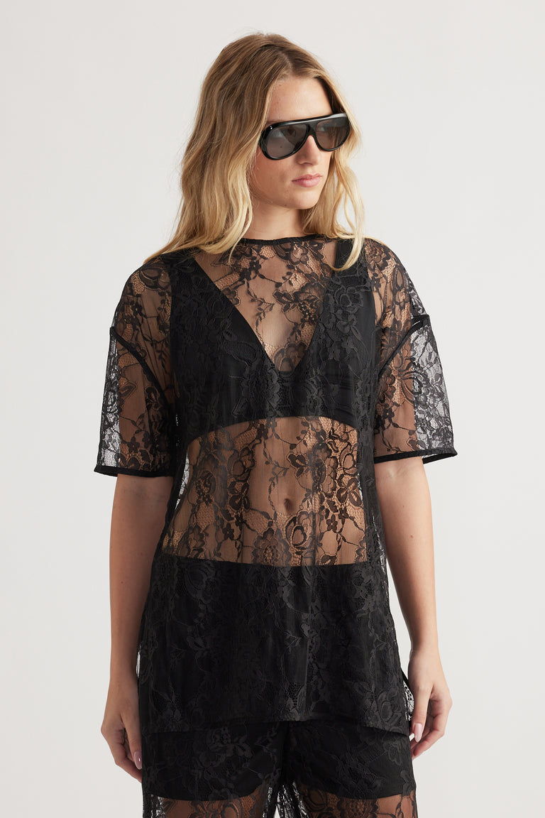 Woman wearing a sheer black lace top with short sleeves and a deep V-neck bralette underneath paired with black shorts