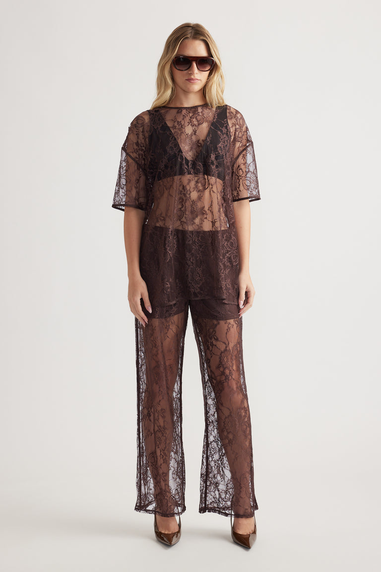 Woman wearing sheer lace top and matching wide-leg pants with dark undergarments and sunglasses on plain background