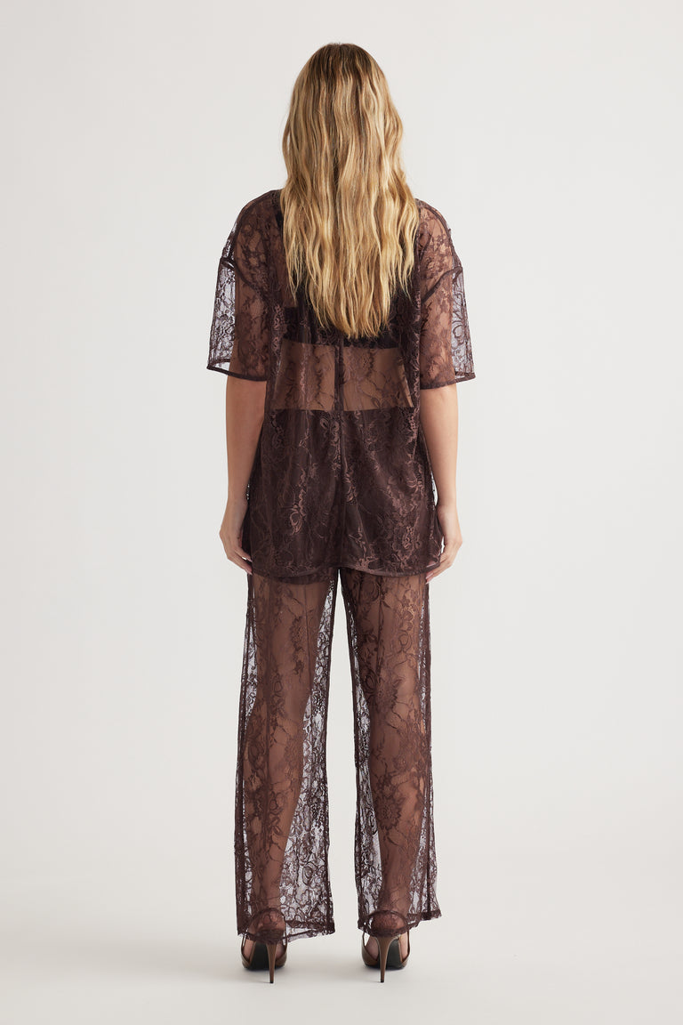 Woman seen from back wearing sheer lace brown top and matching wide-leg lace pants with high heels on plain background