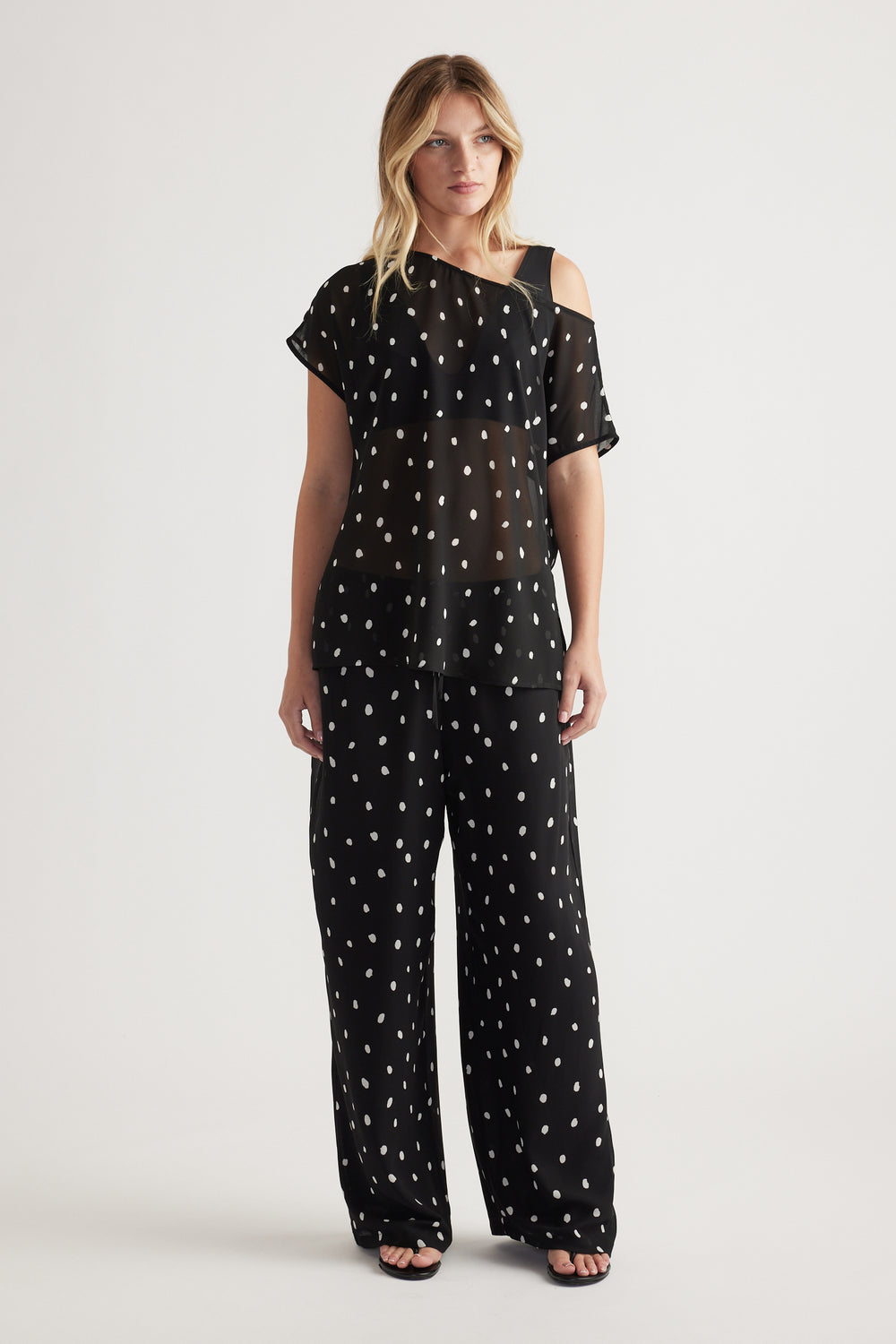 Woman wearing a black sheer off-shoulder top with white polka dots paired with matching wide-leg pants and sandals
