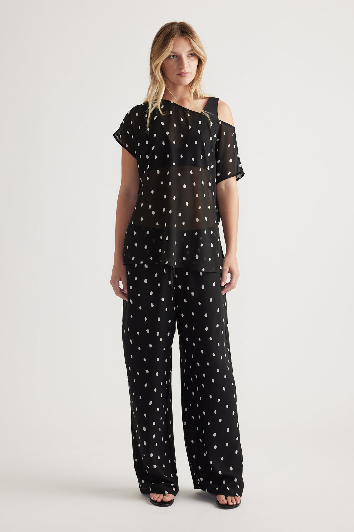 Woman wearing a black sheer off-shoulder top with white polka dots paired with matching wide-leg pants and sandals