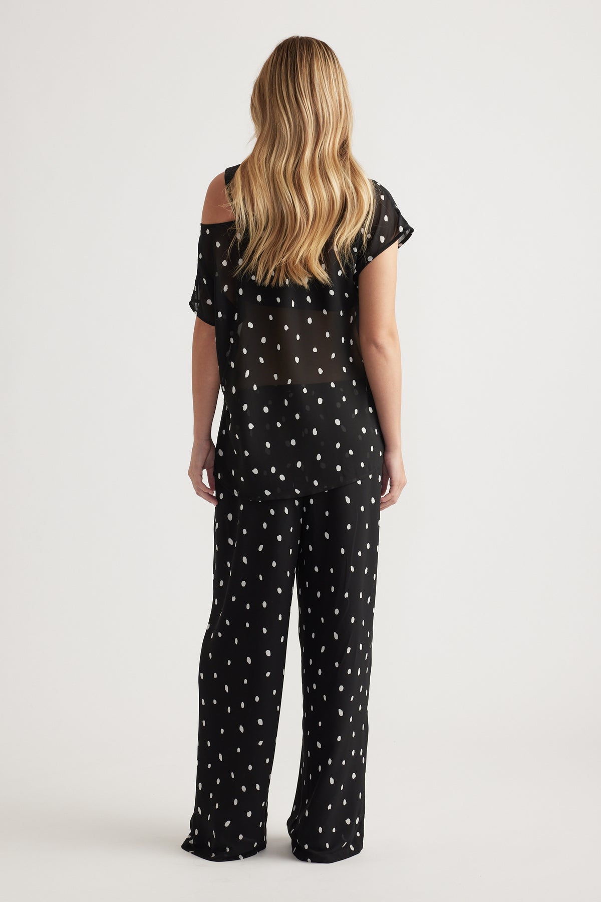 Woman seen from behind wearing a black sheer off-shoulder top with white spots paired with matching loose pants