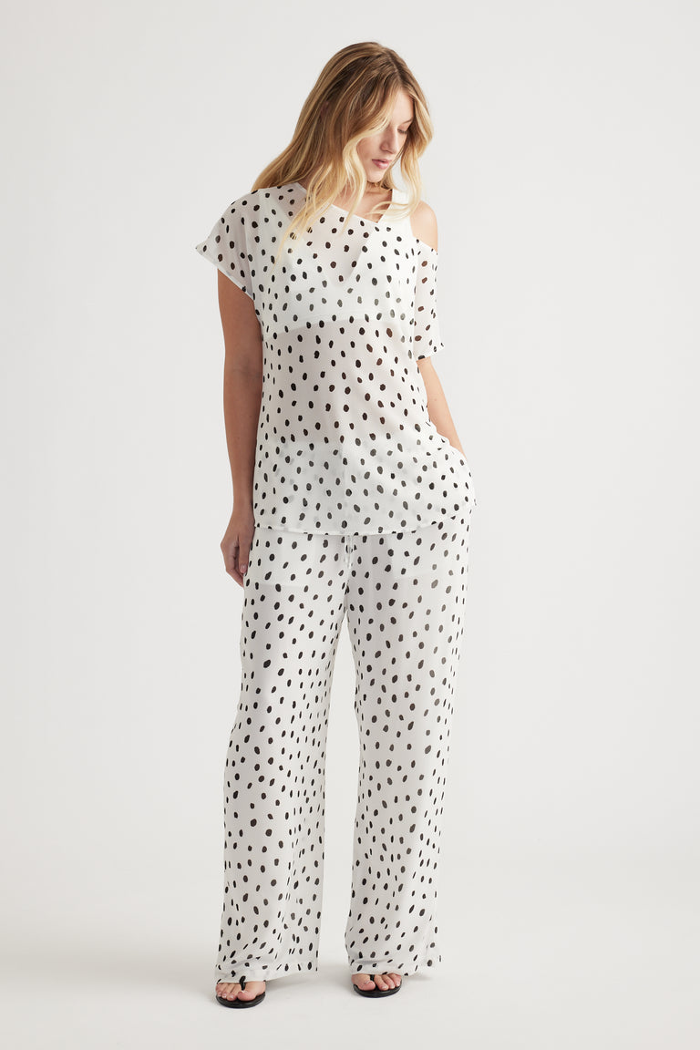 Woman wearing a white off-shoulder sheer top and matching wide-leg pants with a black abstract dot pattern