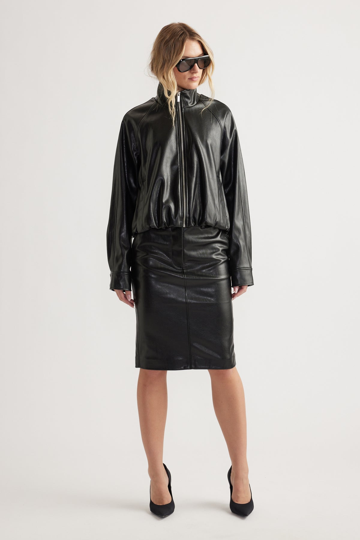Woman wearing relaxed black faux leather bomber jacket with funnel neck paired with fitted black faux leather skirt and heels