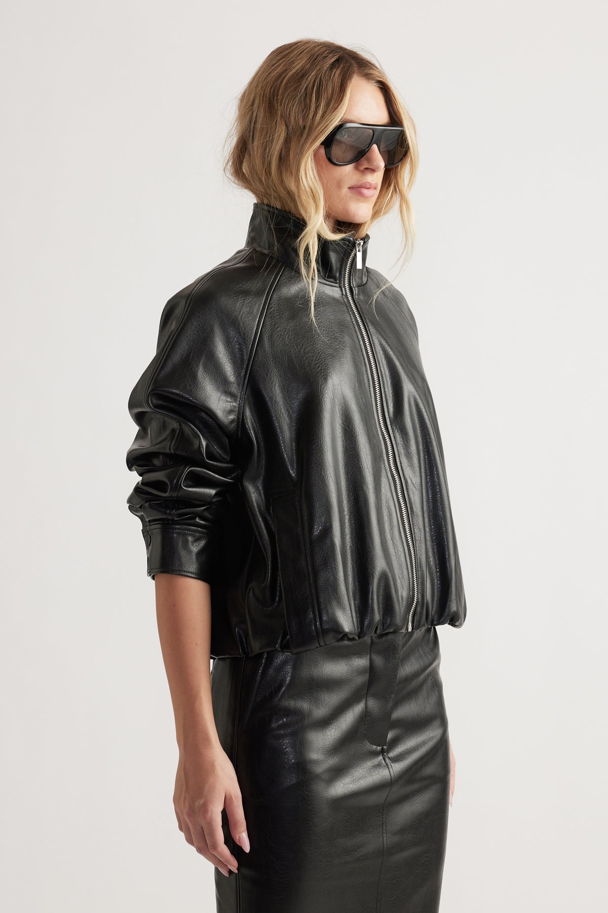Woman wearing black faux leather bomber jacket with zip front and high funnel neck paired with matching skirt and sunglasses