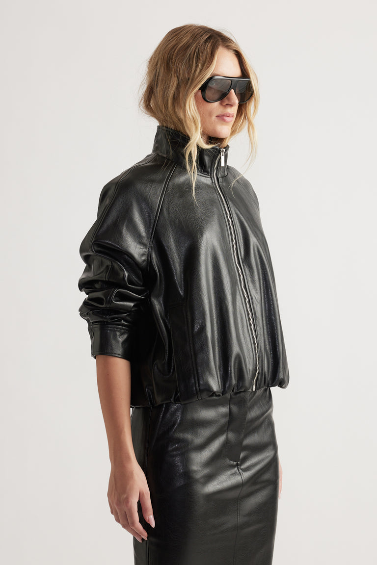 Woman wearing black faux leather bomber jacket with zip front and high funnel neck paired with matching skirt and sunglasses