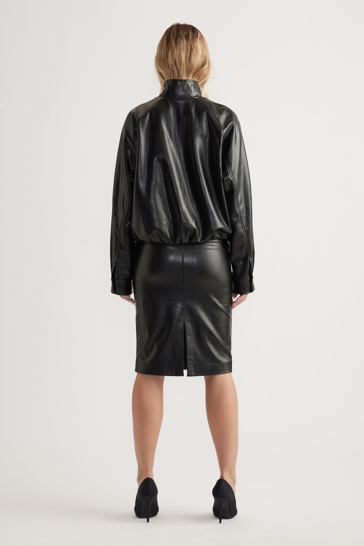 Woman shown from behind wearing a relaxed black faux leather bomber jacket with long sleeves and a knee-length skirt.