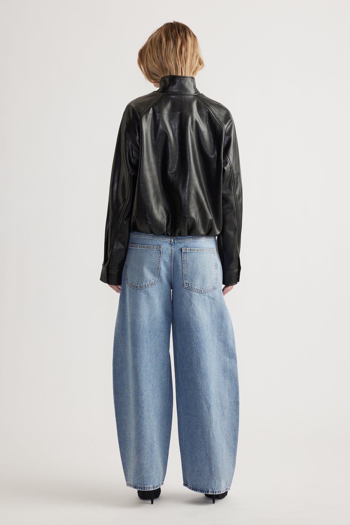 Woman wearing relaxed fit black faux leather bomber jacket paired with wide-leg light blue denim jeans, back view