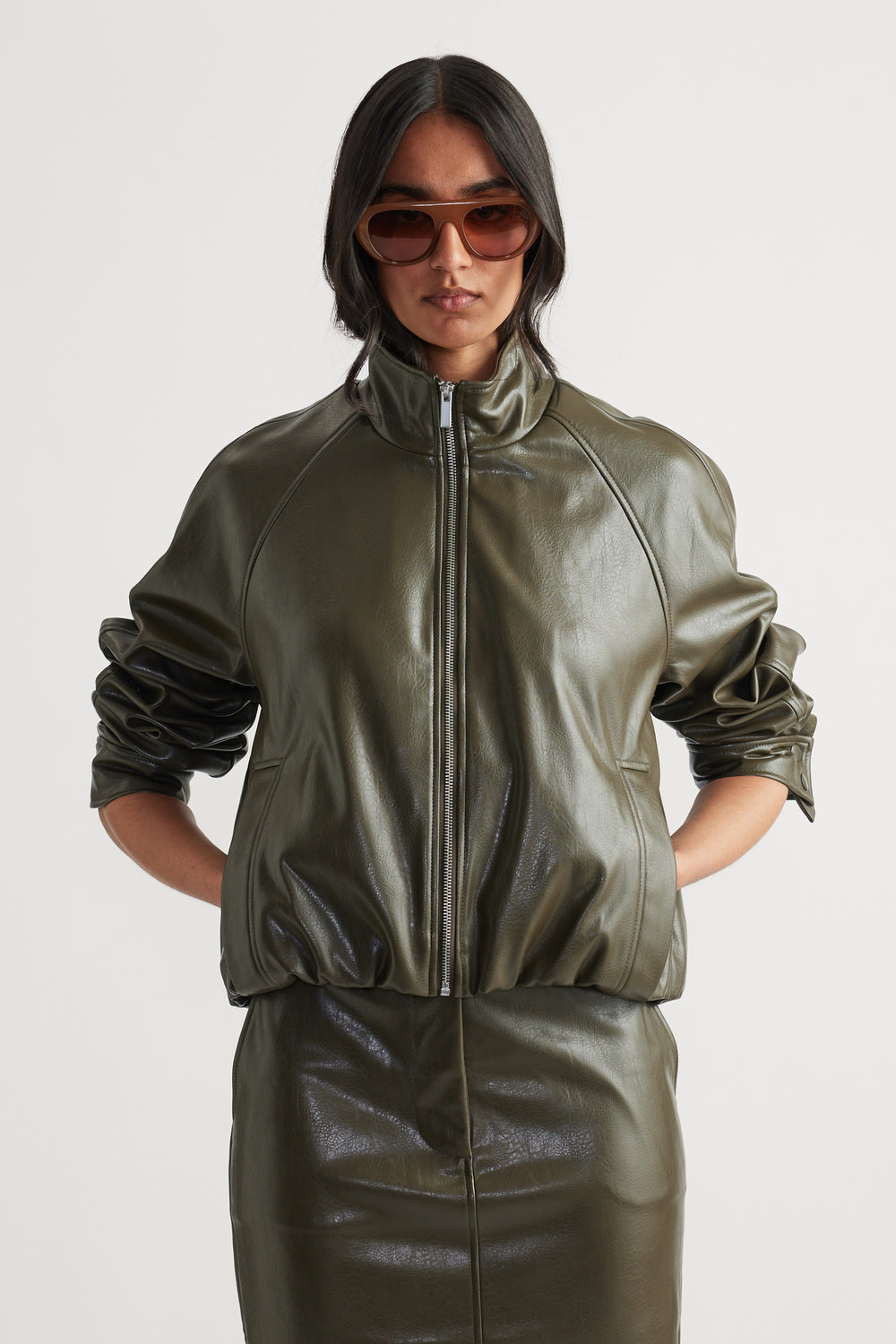 Woman wearing an olive green faux leather bomber jacket with zip closure and ruched sleeves paired with a matching skirt