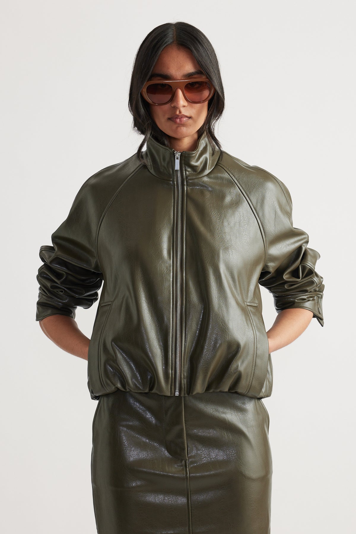 Woman wearing an olive green faux leather bomber jacket with zip closure and ruched sleeves paired with a matching skirt