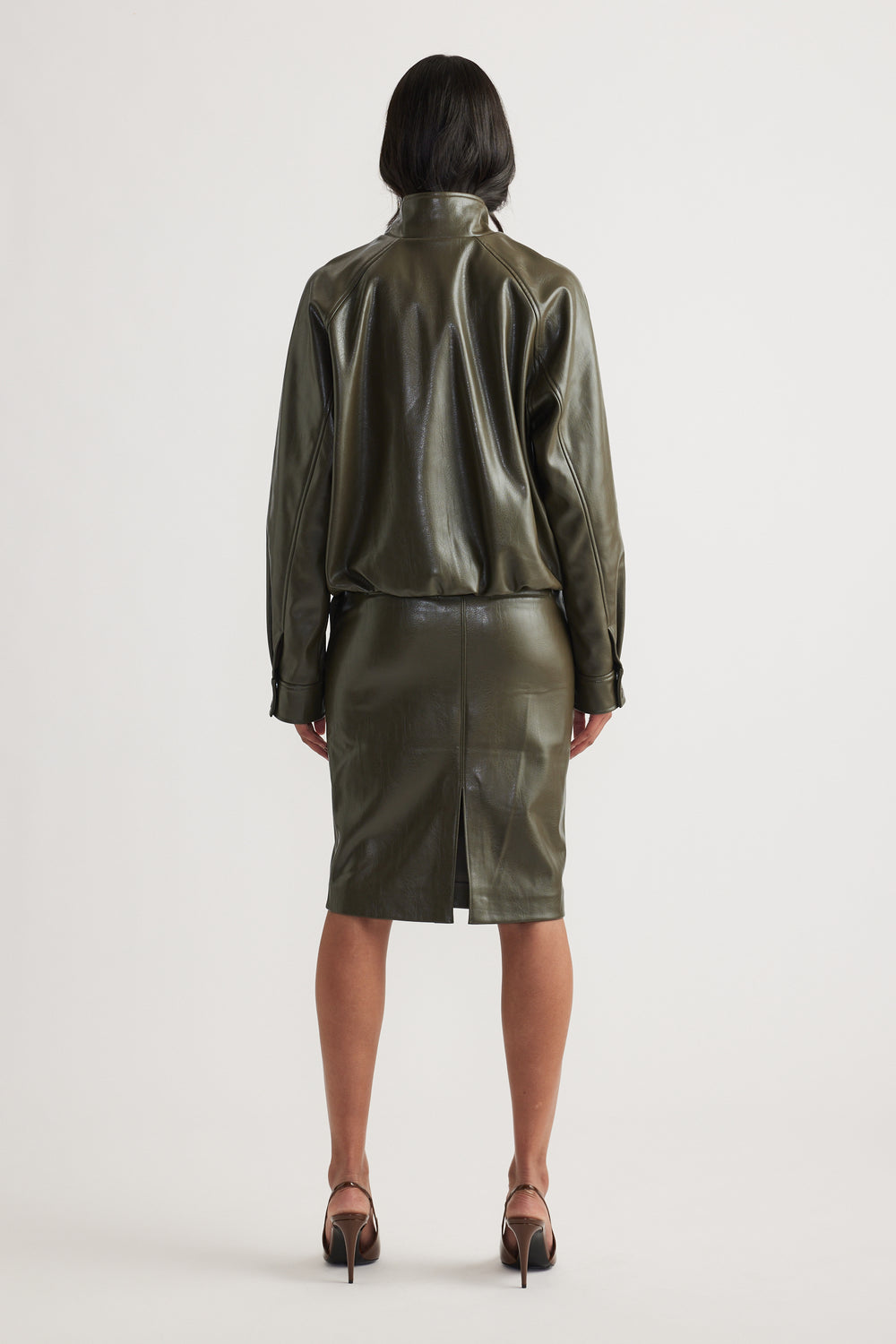 Woman wearing an olive green faux leather bomber jacket with a matching pencil skirt and strappy heels viewed from behind