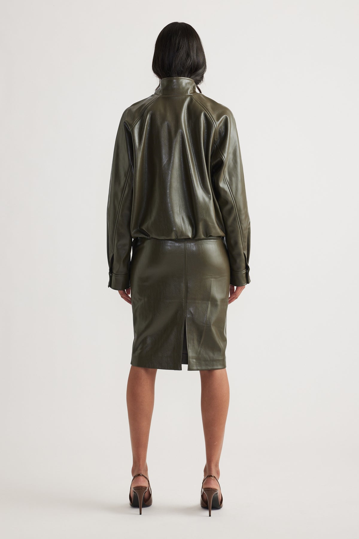 Woman wearing an olive green faux leather bomber jacket with a matching pencil skirt and strappy heels viewed from behind