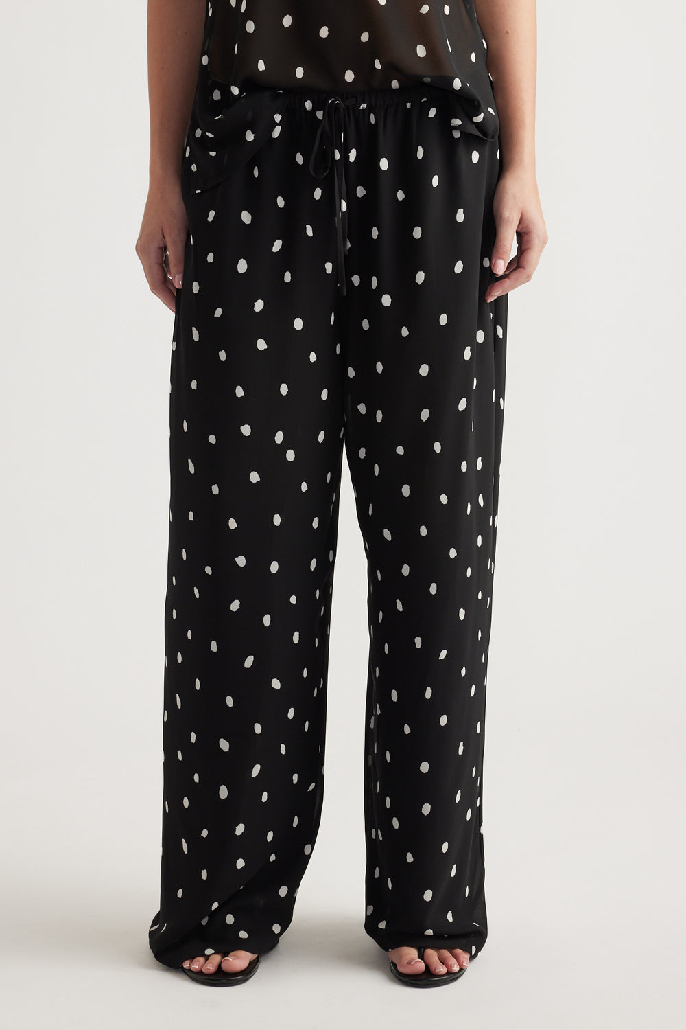 Woman wearing loose black pants with white spots and a drawstring waist paired with black open-toe sandals