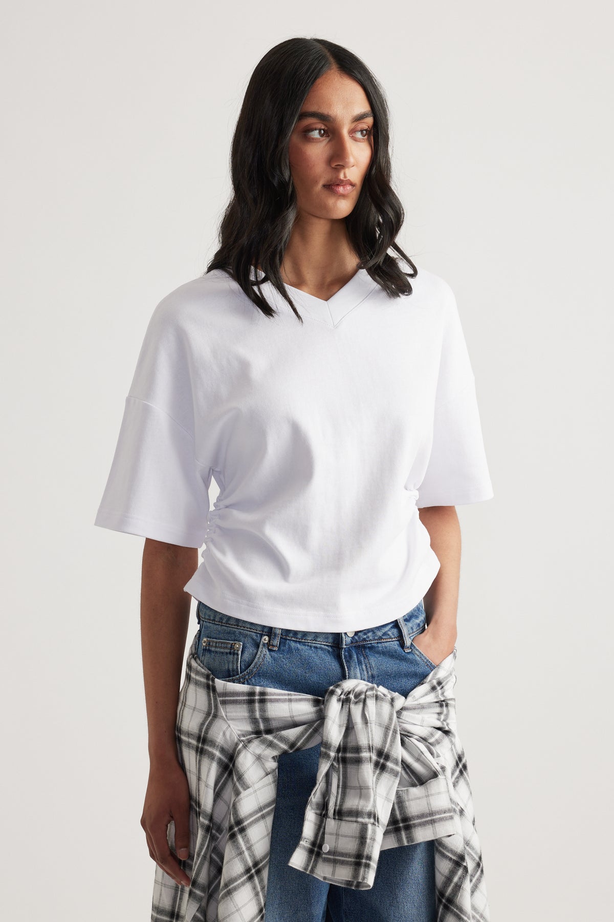 Woman wearing a white boxy V-neck tee with blue jeans and a plaid shirt tied at the waist