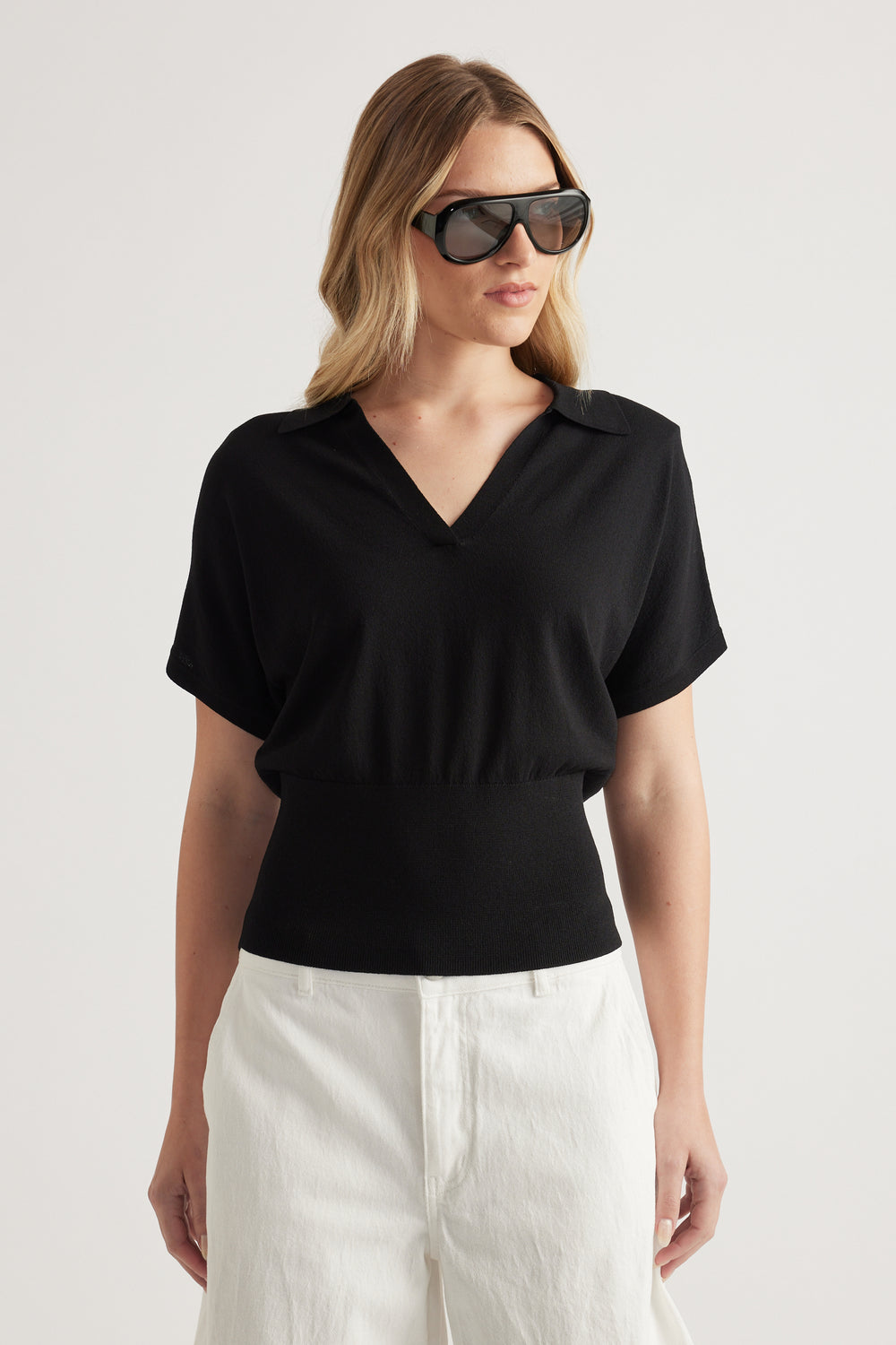Woman wearing a black short sleeve collared knit top with a V neckline and cinched waist, styled with white pants