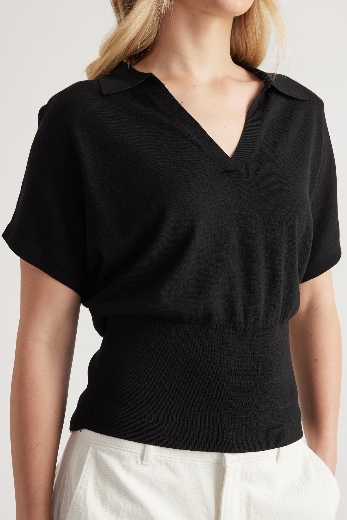 Woman wearing a black short sleeve collared knit top with a cinched waist and white pants